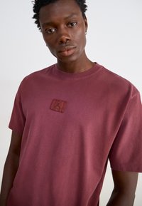 Mørkerød T-shirt lavet af bomuld, med et firkantet mærke med et mørkt logo på brystet. Korte ærmer og rund halsudskæring. Glat tekstur.