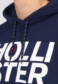 Sudadera azul marino con texto blanco, cuenta con cordones con acentos blancos. Tejido suave, el bolsillo canguro no es visible en el primer plano.