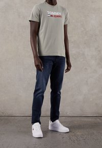 Ljust grå bomullströja med "TOMMY JEANS"-logotyp i röd, marinblå och vit, ihop med mörkblå jeans och vita sneakers på en texturerad bakgrund.