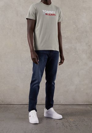 Camiseta de algodón gris claro con el logotipo "TOMMY JEANS" en rojo, azul marino y blanco, combinada con jeans azul oscuro y zapatillas blancas sobre un fondo texturizado.