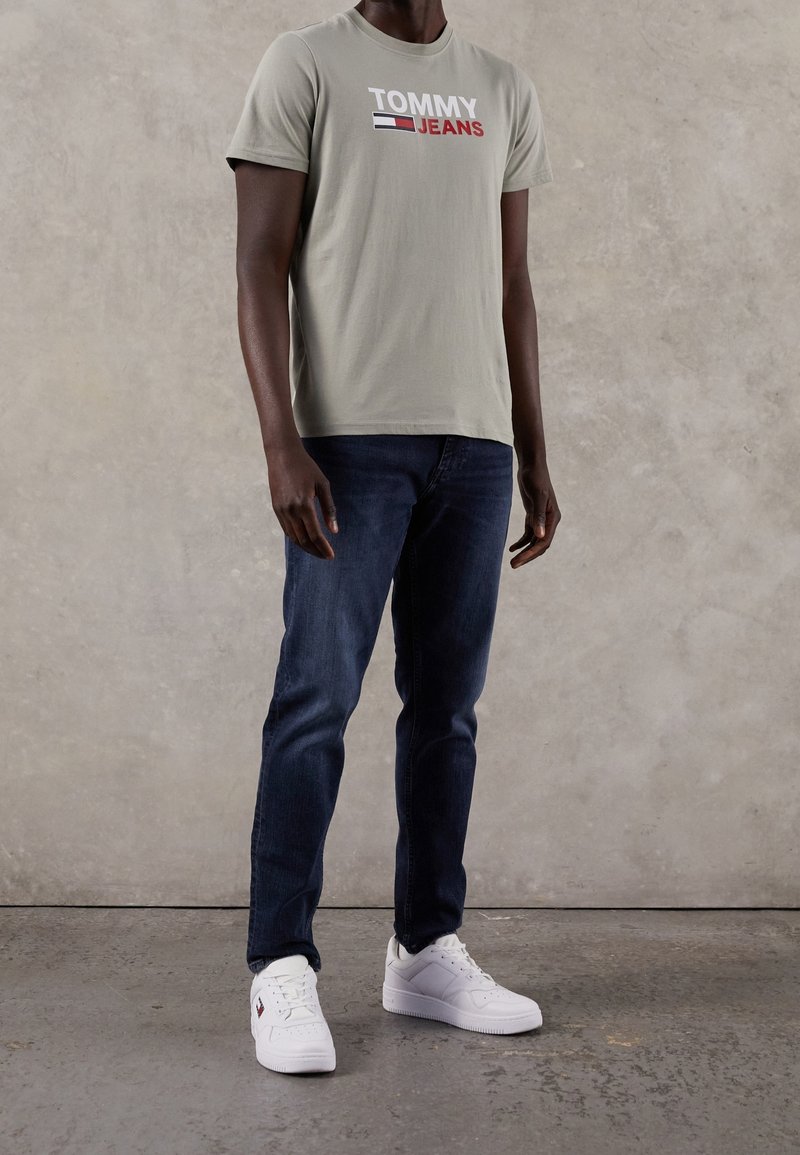 Ljust grå bomullströja med "TOMMY JEANS"-logotyp i röd, marinblå och vit, ihop med mörkblå jeans och vita sneakers på en texturerad bakgrund.