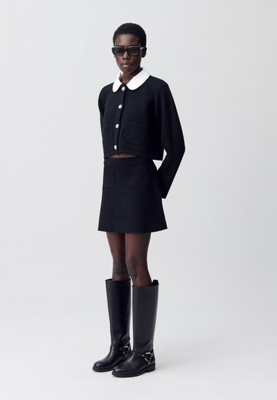 Cardigan noir court avec un col blanc et des boutons dorés, assorti avec une mini-jupe noire. Bottes en cuir noires à hauteur de genou avec des accents en matériel.
