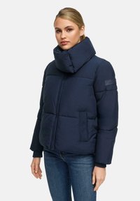 Vrouw in een marineblauwe pufferjack met een oversized kraag, ritsluiting en donkerblauwe jeans, staand tegen een effen lichte achtergrond.