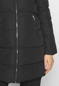 Manteau d'hiver matelassé noir avec fermeture éclair centrale argentée et poche latérale zippée, manche couvrant le poignet, main reposant le long du corps.