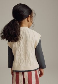 Ärmellose Strickweste in Creme mit Kabelmustern, kombiniert mit einem langärmeligen, dunkelgrauen Hemd und rotgestreiften Hosen. Strukturiert, Rückenansicht.