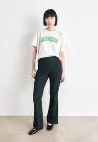 Cremefarvet bomulds T-shirt med "CALVIN KLEIN" logo i grøn, parret med skræddersyede mørkegrønne bukser og sorte flade sko. Minimalistisk design overordnet.