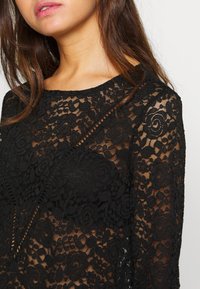 Haut en dentelle noire avec motifs floraux et tourbillons, doté d'un col rond et de manches longues transparentes. La texture est complexe et délicate.