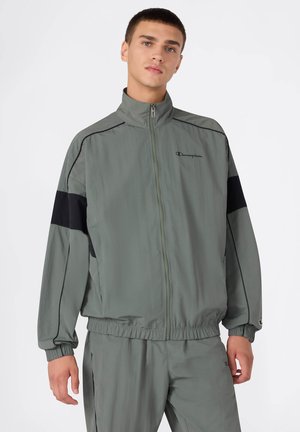 Chaqueta de chándal gris con un alto collar, acentos negros en las mangas, cremallera frontal, puños elásticos y logo en el pecho. Material ligero.