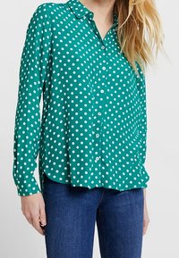 Chemise verte à manches longues avec pois blancs, col, fermeture boutonnée sur le devant, ourlet arrondi, en tissu léger et lisse.