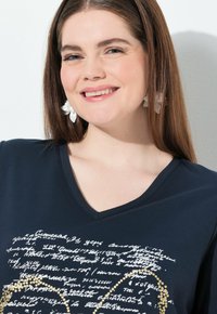 Camiseta de cuello en V en color navy con un patrón de texto manuscrito en blanco y acentos dorados. La tela tiene una textura suave. El modelo lleva pendientes florales.