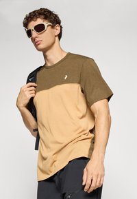 Young man wearing beige and brown color-blocked t-shirt, black shorts with zipper pocket, beige sunglasses, and carrying a black backpack strap.