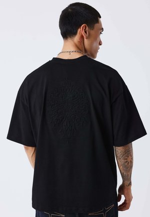 Man met kort zwart haar die een zwarte oversized T-shirt draagt met een verhoogd ingewikkeld kruisontwerp op de rug en een getatoeëerde rechteronderarm.