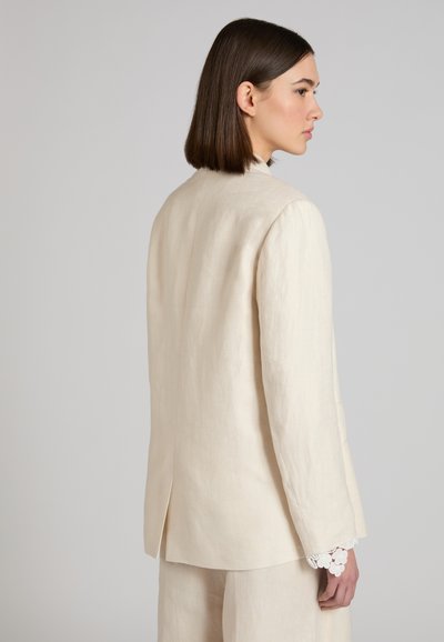 Vrouw met recht bruin haar, gekleed in een crèmekleurig pak met kantafgewerkte mouwen, zijwaarts kijkend tegen een effen grijze achtergrond.