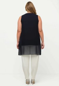 Tunique nera senza maniche con un pannello inferiore trasparente, abbinata a pantaloni bianchi. Il tessuto appare liscio con un design fluido.