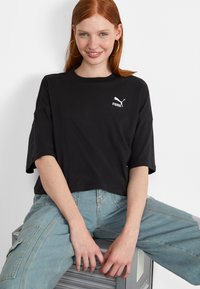 Black cropped t-shirt with a white Puma logo on the chest, paired with light blue high-waisted denim jeans. Smooth texture, relaxed fit.