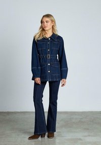 Chaqueta de mezclilla en azul oscuro con cierre de botones, grandes bolsillos y cintura con cinturón. Vaqueros oscuros de campana con costuras visibles. Botas marrones hasta el tobillo.