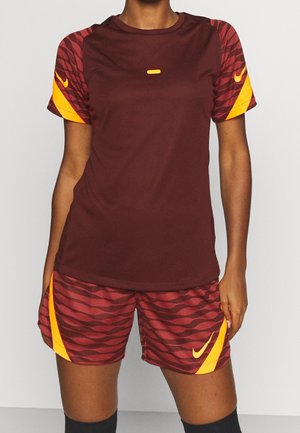 Sportowiec ubrany w bordowo-pomarańczową koszulkę sportową oraz dopasowane, wzorzyste spodenki, stojący z rękami swobodnie opuszczonymi wzdłuż ciała.