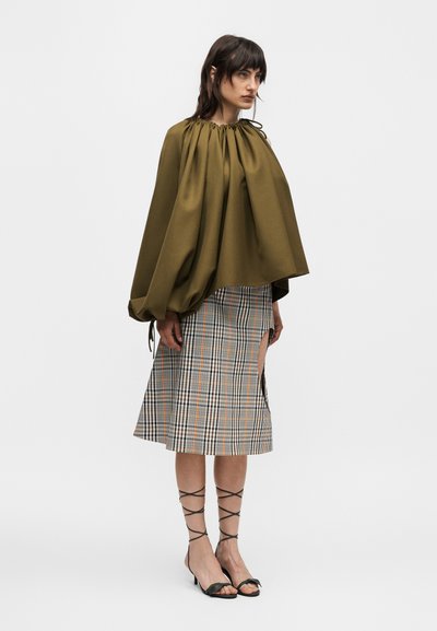 Γυναίκα με oversized τοπ σε ελιά πράσινο με πιέτες, καρό μίντι φούστα με πλαϊνή σχισμή και μαύρα δερμάτινα πέδιλα με κορδόνια και τακούνι, στέκεται σε απλό φόντο.