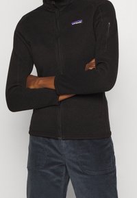 Svart fleecejacka med hög krage och dragkedja. Har en liten, färgglad Patagonia-logotyp på vänster bröst och en ficka med dragkedja på ärmen.