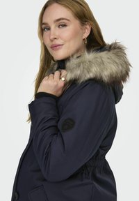 Námornícka parka s kapucňou z umelej kožušiny, elastickým šnúrkovým pásom a logom na rukáve. Hladká textúra a priliehavý strih.