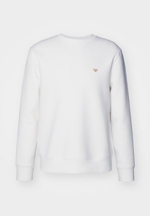 Sweatshirt blanc en tissu doux, avec un col rond et des manches longues. Un petit logo doré est positionné sur le haut de la poitrine gauche.