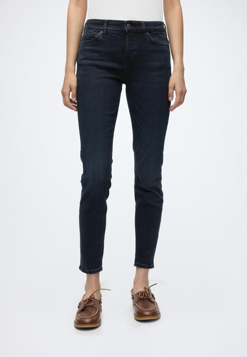 Personne portant un jean slim bleu foncé, un haut blanc et des chaussures en cuir marron à lacets, debout devant un fond clair uni.