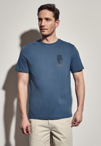 Modrá bavlněná tričko s krátkým rukávem, s grafickým potiskem na levé části hrudníku. Design obsahuje text a geometrický vzor.