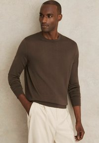 Brauner gestrickter Pullover mit langen Ärmeln, Rundhalsausschnitt und gerippten Bündchen. Kombiniert mit hellen Hosen, vor einem neutralen Hintergrund.