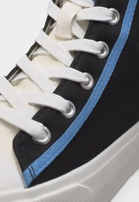 Zbliżenie na czarno-biały tenisówkę z białymi sznurowadłami, srebrnymi przelotkami i niebieskim paskiem wzdłuż boku ponad białą gumową podeszwą.