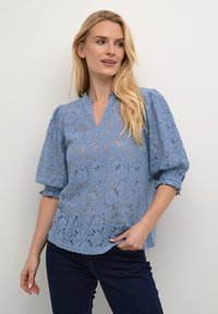 Blusa di pizzo azzurro chiaro con un motivo floreale, maniche a sbuffo corte, scollatura a V e una vestibilità morbida. Abbinata a jeans scuri.