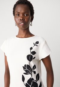 Top bianco con un design floreale nero sul lato sinistro. Il tessuto ha un aspetto texture, con maniche corte a cappuccio e scollatura rotonda.