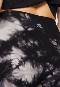 Gros plan d'une personne portant un pantalon à motif tie-dye noir et blanc avec un petit logo floral blanc près de la taille.