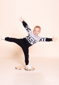 Czarny i szary strój sportowy z graficznym wzorem i elementami kreskówkowymi. Dziecko ma na nogach białe trampki i balansuje na drewnianych krążkach.