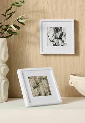 Due cornici quadrate bianche con arte astratta in bianco e nero, un vaso bianco con foglie verdi e una ciotola di ceramica color crema su una superficie in legno.