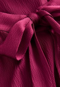 Pinkfuchsia tekstureret stof med et vævet mønster, der har et bundet bælte og draperede folder, som fremhæver materialets dybde og rigdom.