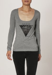 Haut gris à manches longues avec un col en scoop, présentant un graphique triangulaire en sequins noirs et le texte "DEVINE QUI ?" au-dessus.