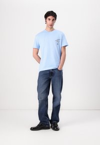 T-shirt en coton bleu clair à manches courtes, col rond, avec un détail de logo noir sur la poitrine, associé à un jean en denim bleu délavé et des chaussures noires.