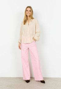 Bézs hímzett blúz V-nyakkivágással és gombos elülső résszel, világos rózsaszín, bő szárú nadrággal és sötétbarna hegyes orrú lapos cipővel párosítva.