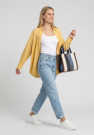 Vrouw die loopt, gekleed in een gele blouse, een wit hemdje, lichtblauwe spijkerbroek, witte sneakers en een gestreepte draagtas met blauwe en witte panelen.