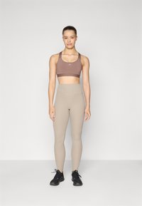 Soutien-gorge de sport marron avec un logo, leggings beiges taille haute avec une texture côtelée, et chaussures de sport noires. Le mannequin se tient devant un fond uni.