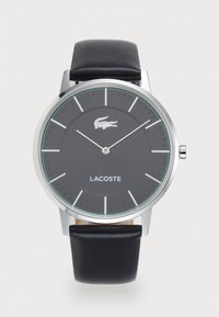 Lacoste klokke med et sølvfarget rustfritt stålhus, svart rund urskive, grønne detaljer, sølvtimeindikatorer og svart lærrem.