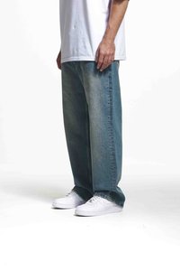 Jeans din denim de un albastru deschis, cu o croială lejeră și o textură uzată, asortați cu adidași albi. Jeansii au un design cu picioare drepte și cusături subtile.