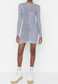 Robe ajustée à manches longues en tissu transparent à carreaux bleu clair et blanc. Portée avec des bottes blanches à lacets et des chaussettes rayées.