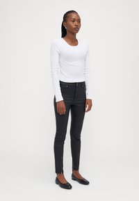 Witte langarm top, gecombineerd met zwarte skinny jeans met ritsdetails bij de enkels. Model draagt zwarte ballerina's en heeft een gevlochten kapsel.