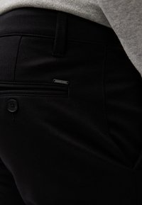 Pantalones negros con una textura suave, que presentan un bolsillo lateral, un cierre de botón y un pequeño logo metálico cerca del bolsillo.