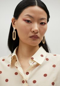 Beige shirt with large red polka dots, collared and buttoned. Dangle earrings in gold and white, model has straight black hair.