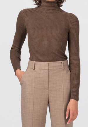 Femme portant un pull à col roulé côtelé marron ajusté et un pantalon beige taille haute, une main dans la poche et un fond neutre.