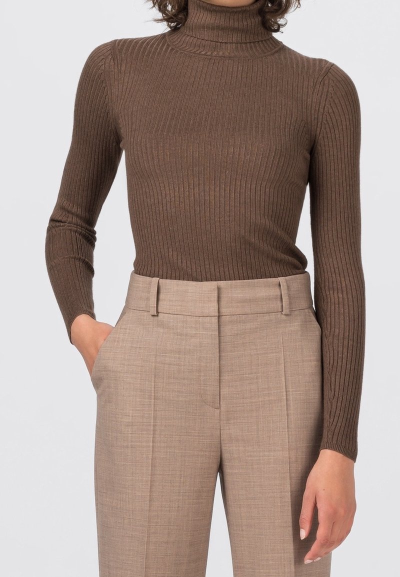 Femme portant un pull à col roulé côtelé marron ajusté et un pantalon beige taille haute, une main dans la poche et un fond neutre.