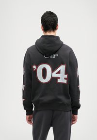 Čierna mikina s kapucňou, na chrbte s veľkým číslom '04' v striebornej farbe s červeným okrajom, so vzormi hviezd na rukávoch. Materiál vyzerá mäkký a textúrovaný.