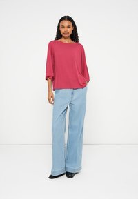 Top solto vermelho com mangas largas, combinado com jeans de perna larga azul claro. Modelo está de pé contra um fundo branco, usando sapatos pretos.
